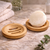 Round bamboo soap dish with drainage slots holding a solid shampoo bar on a bathroom countertop with towel in the background