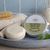 Shea Butter and Ground Oats shampoo bar displayed beside its tin packaging on a blue ceramic tray in a soft bathroom setting, with pale blue tiles, folded towel, wooden brush and diffuser in the background.