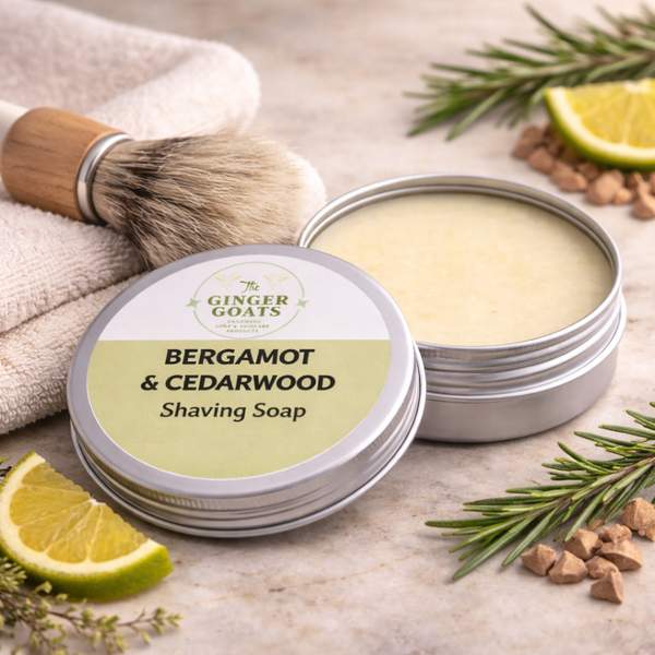 Bergamot and cedarwood goat milk shaving soap in a metal tin with lid, styled with a shaving brush, towel, and natural citrus and herb elements in a bathroom setting.