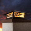 A classic, traditional sloped-roof column mounted pier light fixture with crown molding accent, a bronze finish, floral artwork and golden glass. In the background is a dramatic, cloudy sunset. 