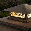 A classic, traditional sloped-roof column mounted pier light fixture with crown molding accent, an aged bronze finish, floral artwork and golden glass. In the background is the garden where the lantern is installed. 