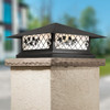 A classic, traditional sloped-roof column mounted pier light fixture with crown molding accent, a black finish and white glass. The lantern is installed on a driveway in front of two separate garages of a large house. 