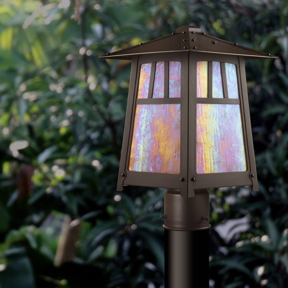 A classic Arts & Crafts style post mount light fixture with an aged bronze finish and iridescent glass in an overgrown garden.