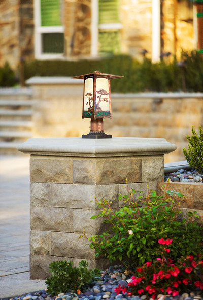 A rustic, artistic post mounted landscape light fixture with Japanese Maple artwork, a raw copper finish and off white glass installed on a stone block pillar in a driveway.