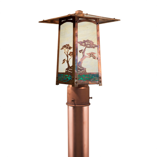 A rustic, artistic post mounted lantern with a peaked roof, Monterey Cypress artwork, a raw copper finish and creamy glass.