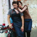 Man and woman wearing dark indigo denim bib aprons with brown leather straps in an industrial outdoor setting
