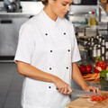 Woman in white short sleeve chef jacket with black stud buttons, chopping vegetables in a professional kitchen