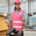 Male worker wearing a pink high-visibility safety vest and matching hard hat, standing in a warehouse while operating a pallet jack. He is also dressed in a grey t-shirt and navy work pants with reflective stripes.