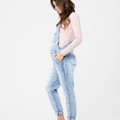Model wearing pale blue denim maternity overalls with adjustable straps, chest pocket, and rolled cuffs