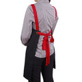 Back view of black cotton canvas bib apron with contrast red cross-back straps and tie, worn over houndstooth shirt