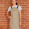 Woman wearing khaki cotton canvas bib apron with black cross-back straps, brass snap buttons, and front patch pocket