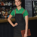 Black denim bib apron with tan leather straps and chest pocket worn over emerald green polo shirt