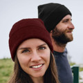 Woman in deep red ribbed knit beanie with cuffed brim, man in black beanie behind her outdoors
