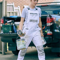 White cotton drill bib overalls with black contrast straps and multiple utility pockets, worn by painter holding paint supplies