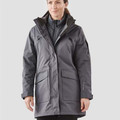 Woman wearing charcoal grey long waterproof system jacket with black inner vest, zip chest pocket and large flap hip pockets