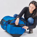 Woman opening a large royal blue and black 85L duffel bag with reinforced straps and zip panels on a grey background