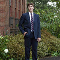 Young male model wearing navy formal school trousers with belt loops, paired with navy blazer and striped tie outdoors