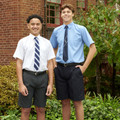 Two students wearing formal short sleeve school shirts in white and pale blue with ties and dark grey shorts outdoors