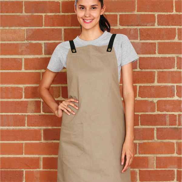 Woman wearing khaki cotton canvas bib apron with black cross-back straps, brass snap buttons, and front patch pocket