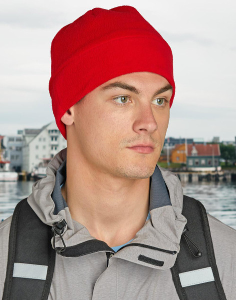 Man wearing a red polar fleece beanie outdoors near waterfront in grey technical jacket