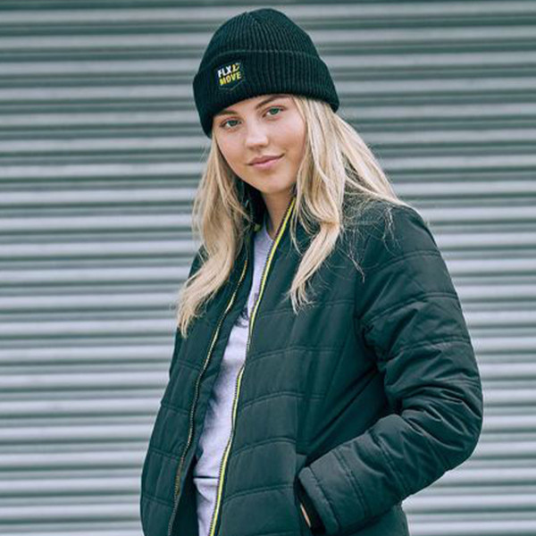 Female model wearing black ribbed cuffed beanie with small embroidered logo patch, styled with black quilted jacket