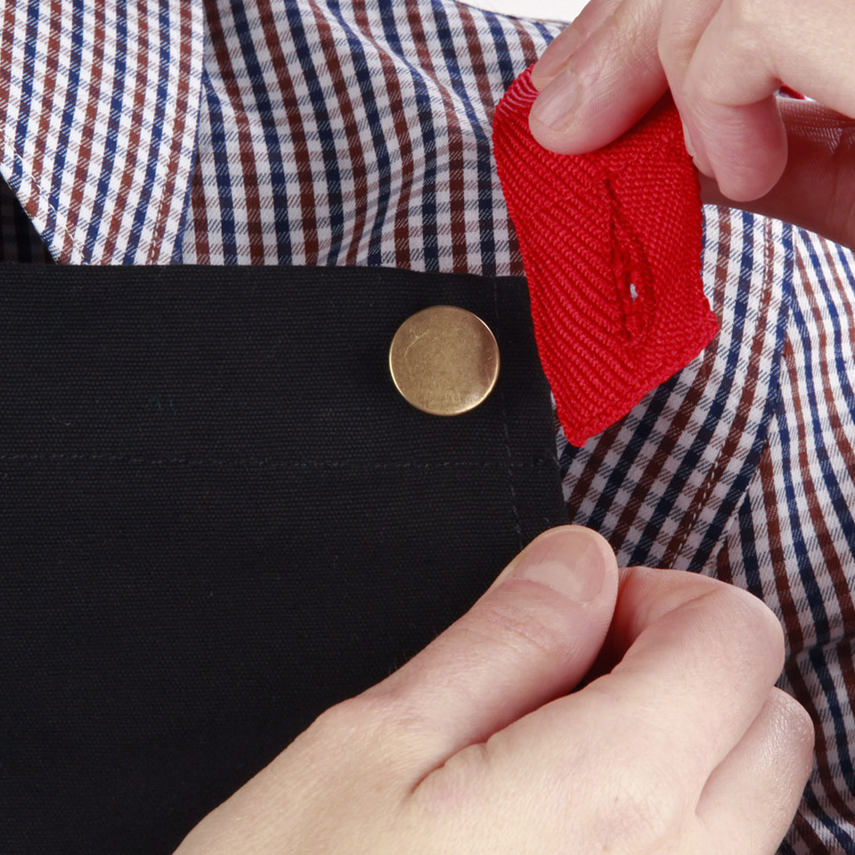 Black cotton canvas bib apron with brass rivet detail and red webbing ties shown against gingham shirt