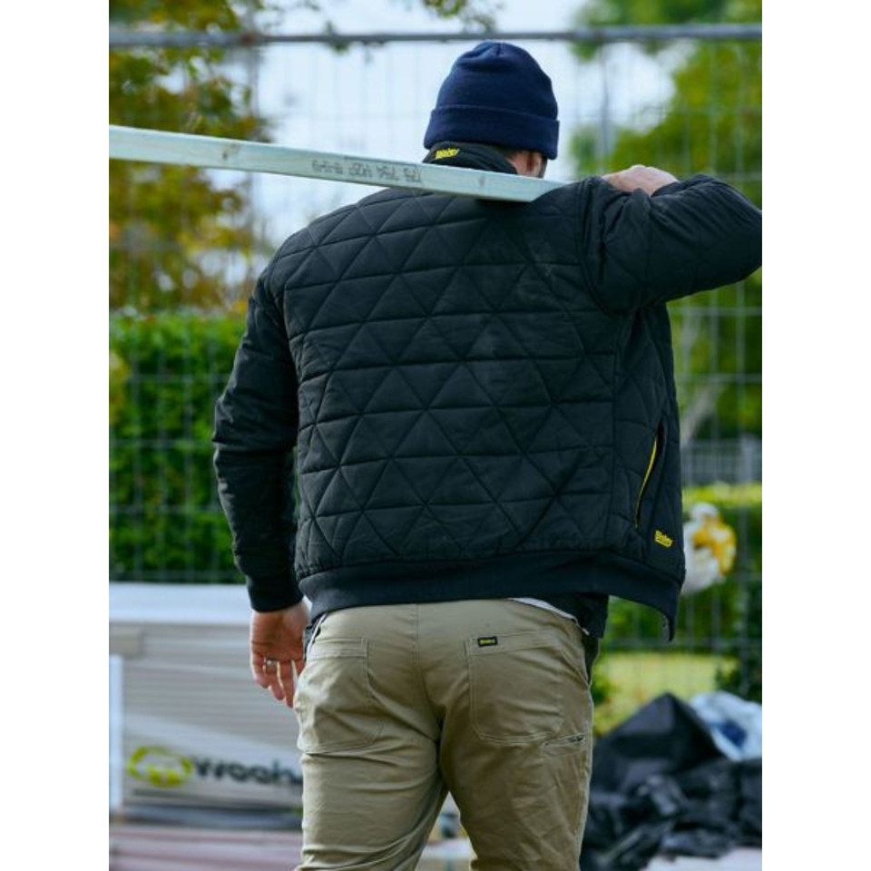 Man on worksite carrying metal beam, wearing black diamond quilted bomber jacket with yellow zip detail, rear view