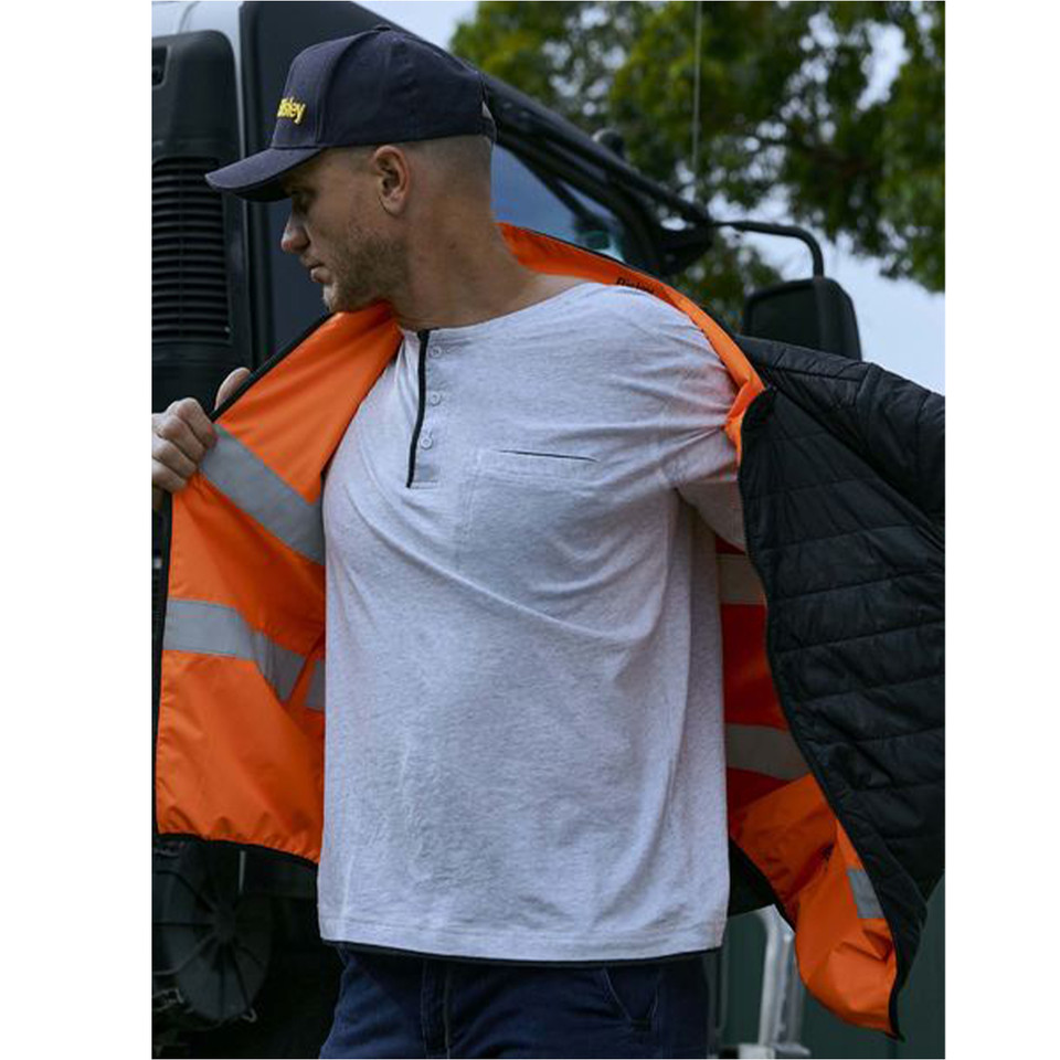Man opening reversible hi vis jacket showing orange interior with reflective tape and quilted black exterior, near truck