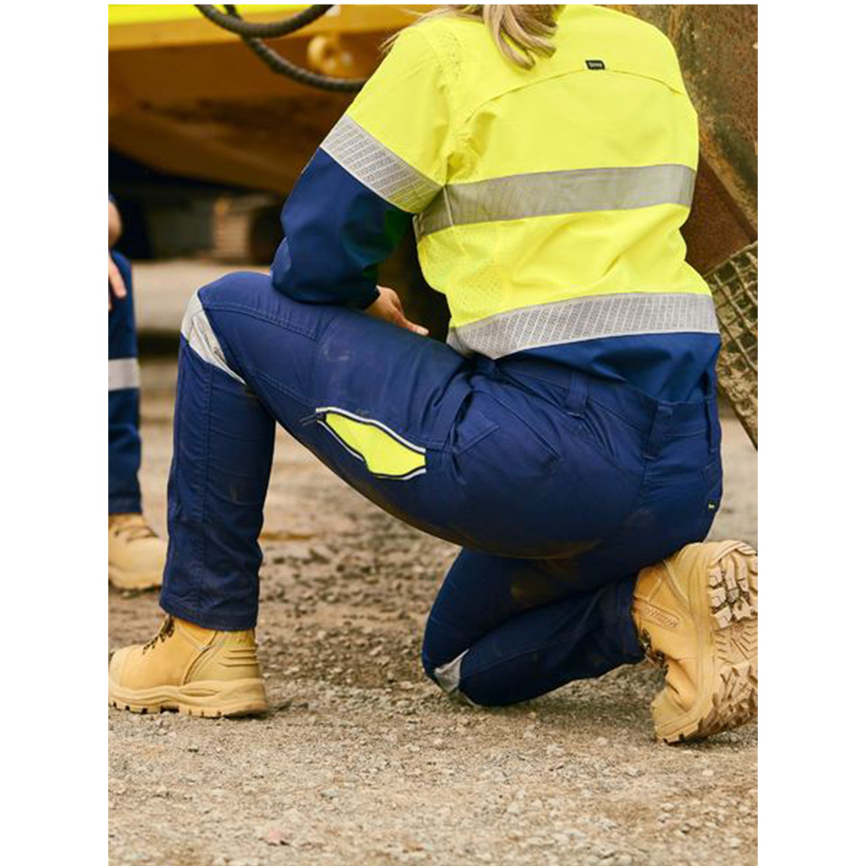 Woman kneeling on worksite in navy stretch ripstop cargo pants with yellow hi-vis knee patch detail and tan work boots