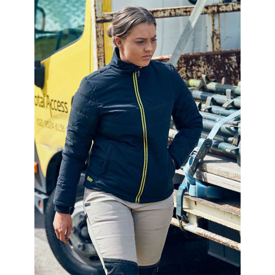 Woman in navy quilted puffer jacket with yellow zip trim, worn near a work vehicle in a trade site setting