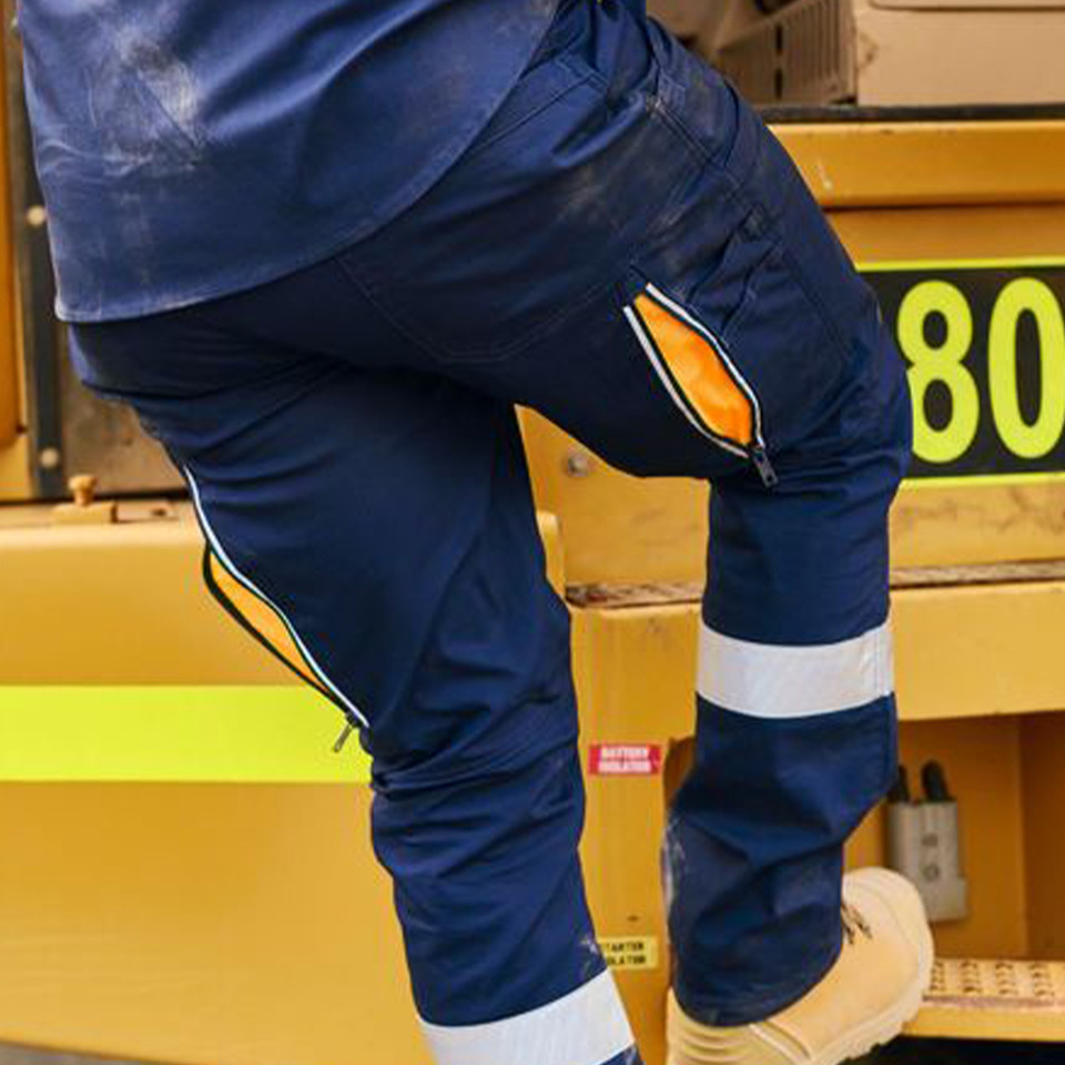 Navy stretch cargo work pant with orange-lined knee vent zip and silver reflective ankle tape, worn on industrial steps
