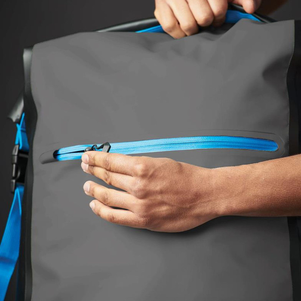 Hand opening cyan zip pocket on charcoal waterproof roll-top backpack with cobalt blue shoulder strap