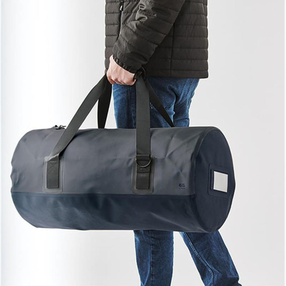 Man carrying large navy blue 65L barrel duffel bag with black webbing handles and welded seam construction