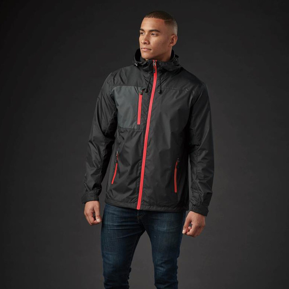 Man wearing black waterproof shell jacket with charcoal chest panel, red contrast zip and side pockets, adjustable hood