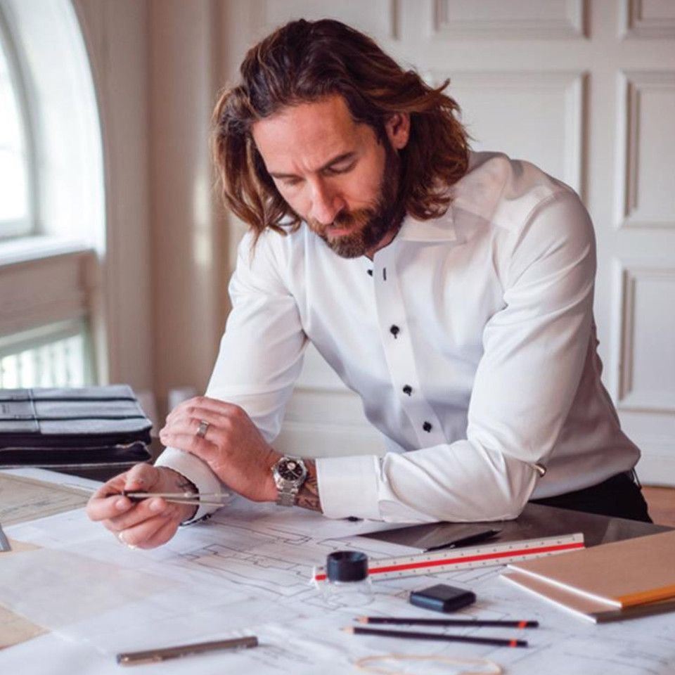 Man in white slim-fit dress shirt with dark contrast buttons working at a drafting desk with blueprints