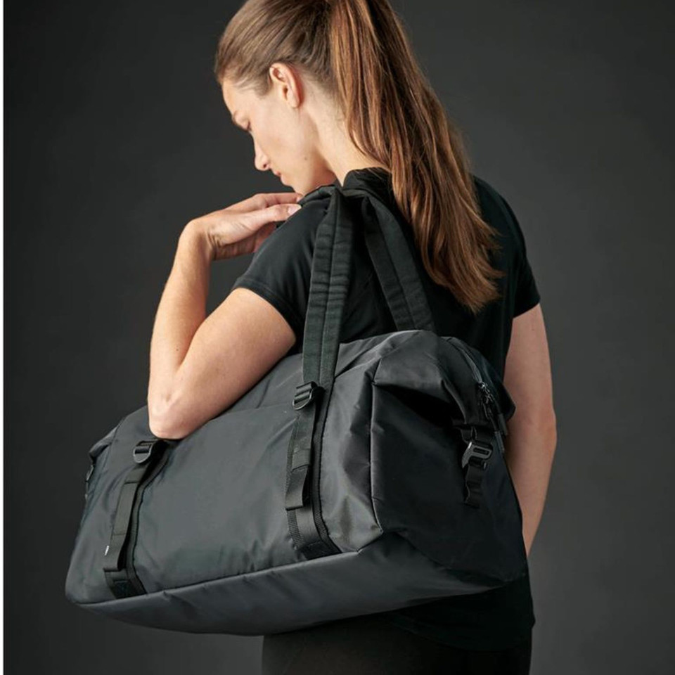 Woman carrying a charcoal grey 32L nylon duffle bag over shoulder with buckle straps and dual carry handles