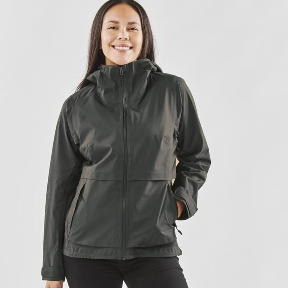 Woman wearing a charcoal technical shell jacket with attached hood, centre-front zip, and zippered hand pockets