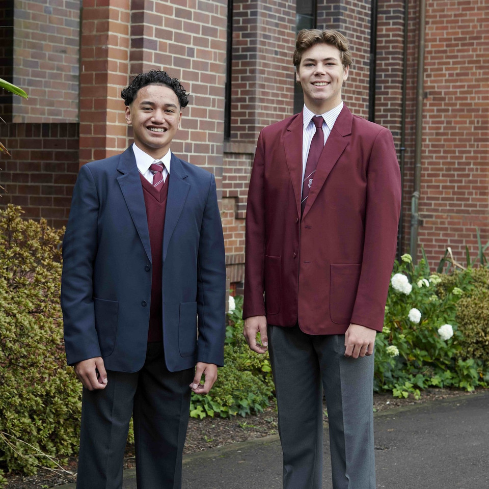 Two male students wearing formal school blazers — navy and burgundy — with white shirts and ties, outdoors near brick building