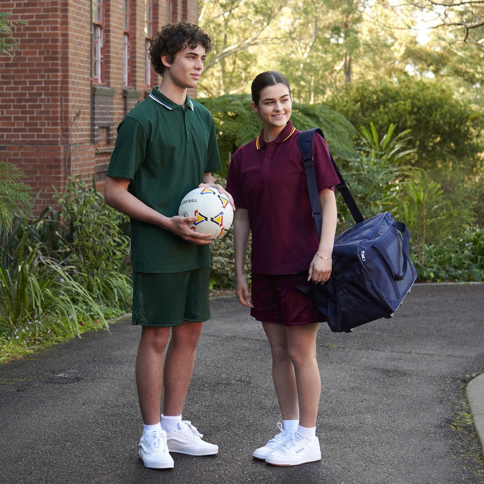 Two students in bottle green and maroon twin stripe polo shirts with matching shorts in a school courtyard