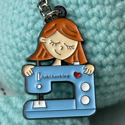 Front view of the Juki Junkies key chain featuring the signature logo girl hugging her sewing machine, designed by Gigi, measuring approximately one and three-quarter inches by one and one-quarter inches
