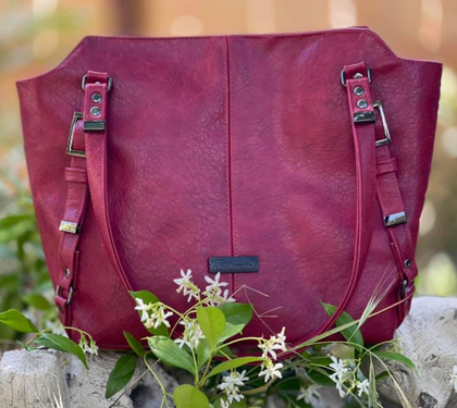 Burgundy leather handbag with adjustable straps displayed outdoors among small white flowers.