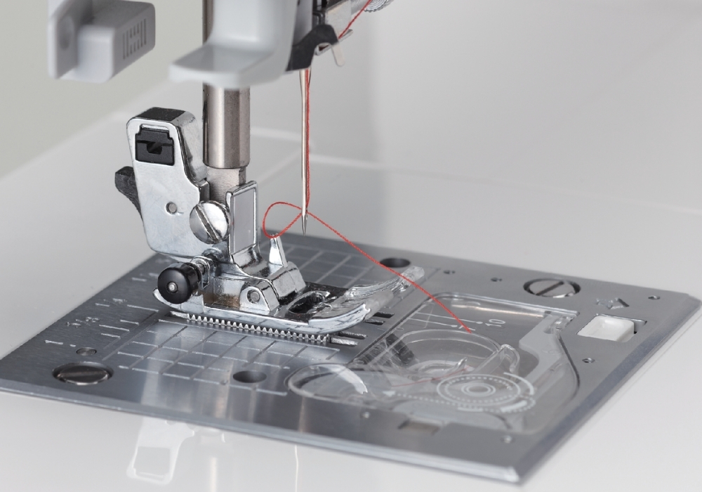 Close-up of a sewing machine needle threaded with red thread, positioned over the needle plate.