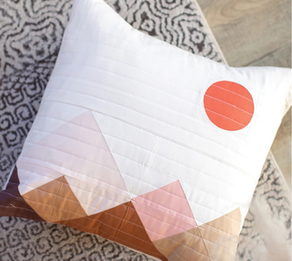Quilted throw pillow with a mountain and sun design in pink, orange, and tan resting on a patterned rug.
