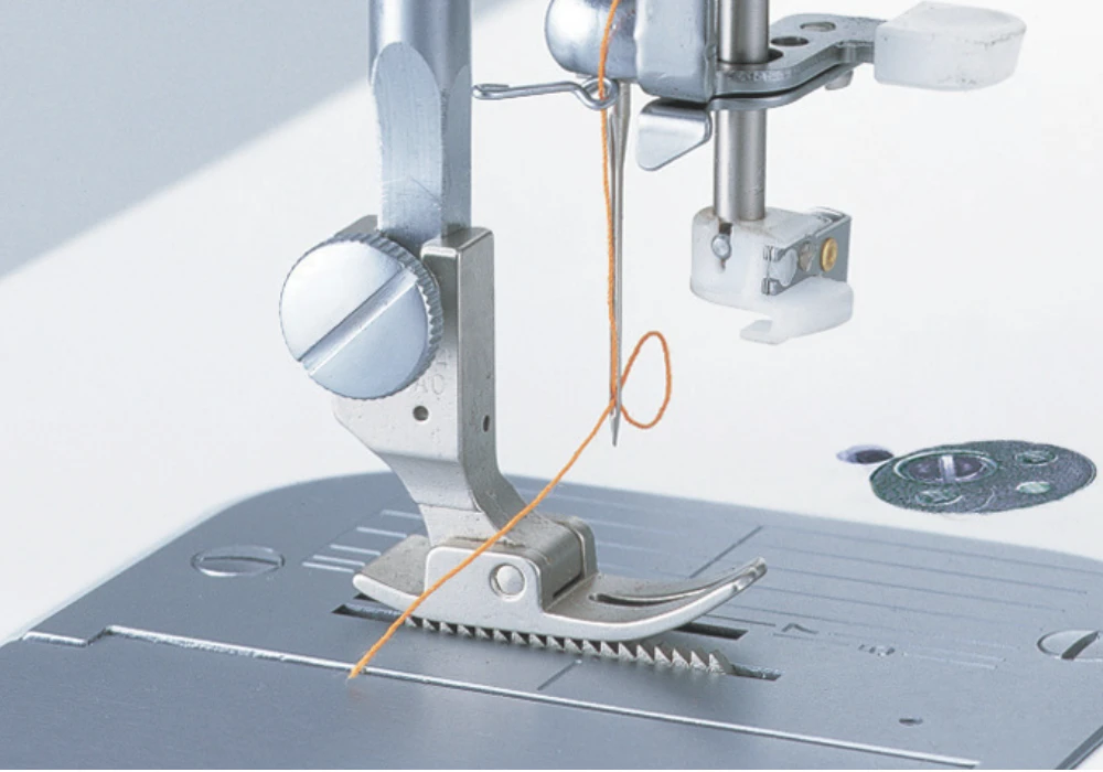 Close-up of a sewing machine needle and presser foot with orange thread, positioned over the feed dogs.