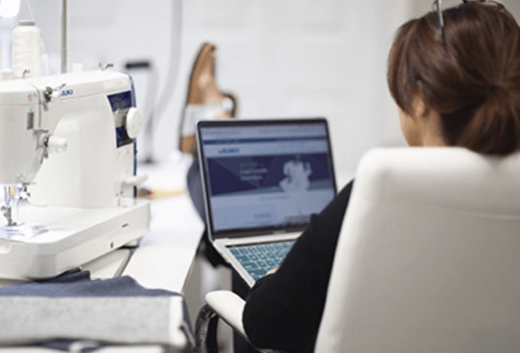 Back view of Gigi working on a laptop in front of a Juki sewing machine.