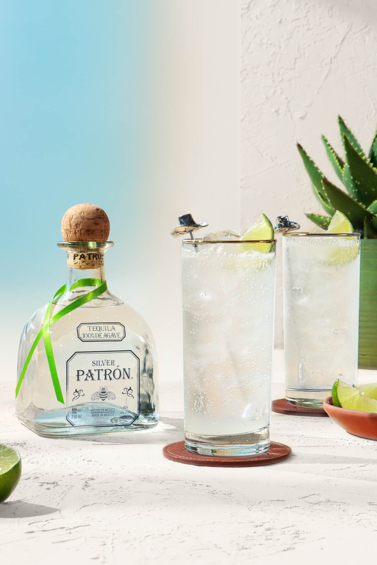 A bottle of PATRÓN Silver and 2 glasses of PATRÓN Ranch Water cocktail with cowboy hat stir rods on leather coasters.