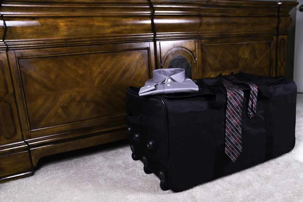 Black Buffalo Duffel in bedroom on side showing foot locker profile & rigid structure with clothes on top of it.