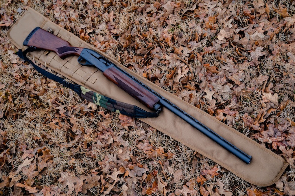 A Coyote Brown Shotgun Scabbard on the ground with brown leaves with a brown and black rifle with camo and black strap laid on top of it.