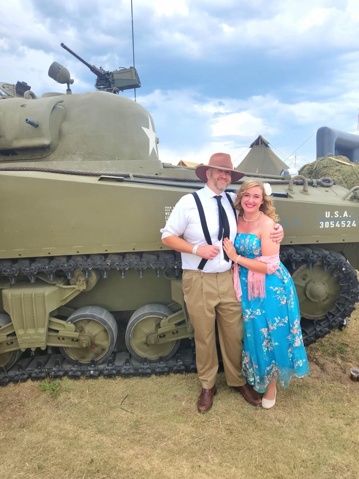 Woman in a cream floral lace A-line dress standing with a gentleman beside a vintage military tank
