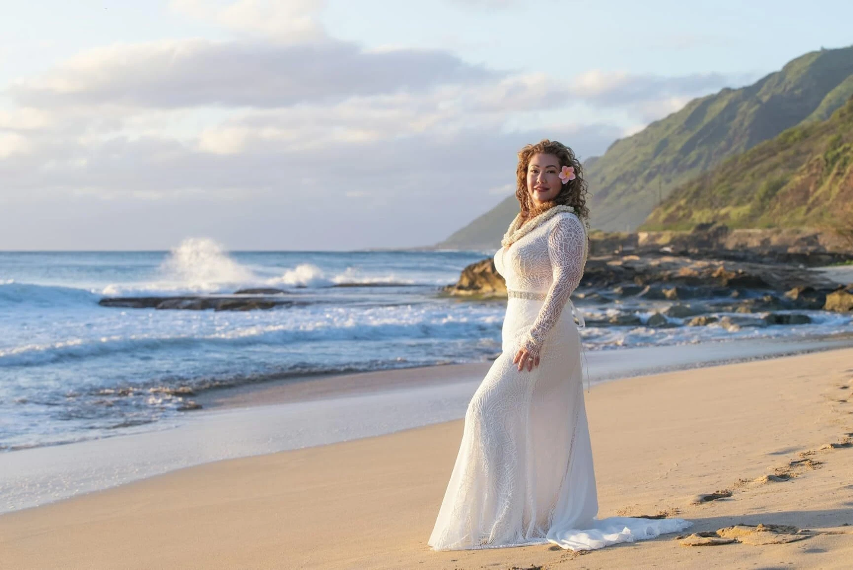 Bride wearing a lace wedding gown on an Oahu beach with mountains behind her